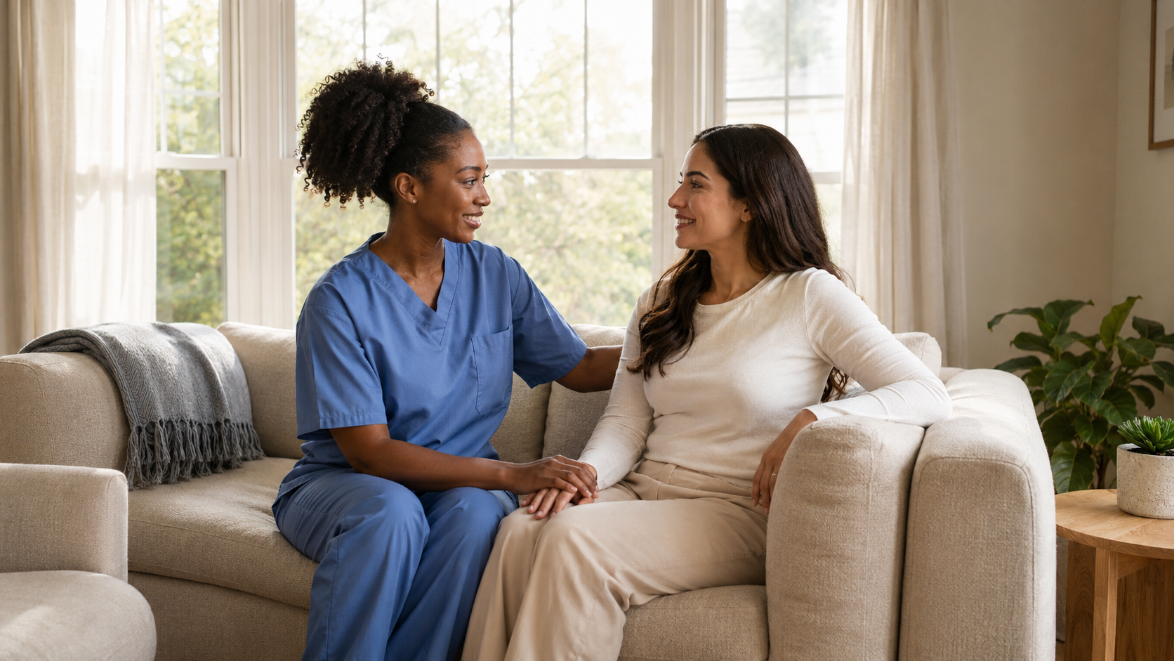 A caregiver in blue scrubs speaking warmly with an older woman in her home